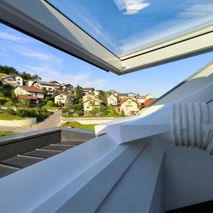 May include: A modern skylight with a clear glass panel, offering a view of a blue sky and a neighborhood of houses. The skylight is framed in white, with a white architectural element in the foreground.