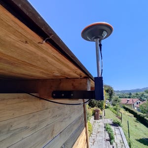 May include: A GPS receiver fixed to a wooden structure. The receiver features a grey and orange dome, connected to a black bracket. The wooden structure is constructed from light brown planks, set against a clear blue sky.