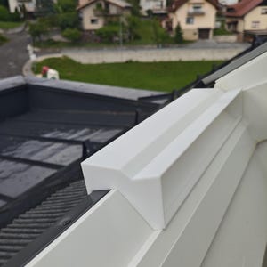 May include: A white, V-shaped plastic component mounted onto the edge of a window frame. The piece features a smooth, matte finish with a distinct angular profile designed to fit securely over the top of the window structure.