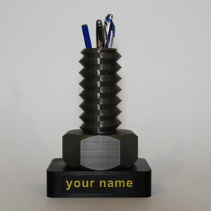 Puede incluir: Un organizador de escritorio de color negro impreso en 3D con forma de un gran tornillo con una base hexagonal. El organizador tiene un espacio para bolígrafos y lápices. La base tiene el texto "your name" impreso en dorado.