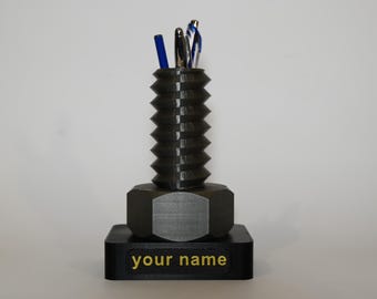 May include: A black 3D printed desk organizer shaped like a large bolt with a hexagonal nut base. The organizer has a space for pens and pencils. The base has the text "your name" printed in gold.