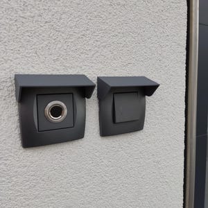 Può includere: Due scatole elettriche da parete esterne nere con coperchi. La scatola a sinistra ha un pulsante del campanello rotondo. La scatola a destra ha un interruttore della luce.