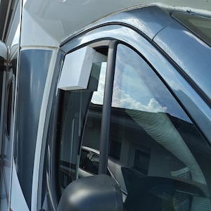 May include: A white rectangular air vent deflector mounted on the upper frame of a vehicle window. The accessory is positioned near the side mirror of a white and dark gray camper van, featuring a clean, solid surface and a simple geometric design.