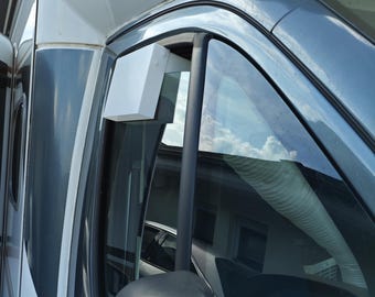 May include: A white rectangular air vent deflector mounted on the upper frame of a vehicle window. The accessory is positioned near the side mirror of a white and dark gray camper van, featuring a clean, solid surface and a simple geometric design.