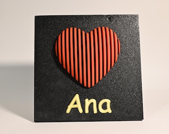 May include: A black square plaque with a red heart with black stripes and the name "Ana" written in yellow.