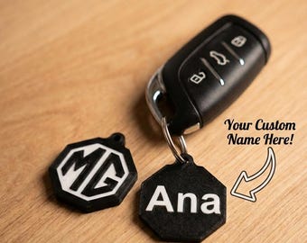 May include: Black car key with a custom keychain that says "Ana". The keychain is black with white lettering. A second black and white MG logo keychain is also present. The key and keychains are on a wooden surface.