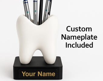 May include: A white tooth-shaped pen holder with a black base. The base has gold lettering that reads "Your Name". The pen holder contains three silver pens and one black pen. The text "Custom Nameplate Included" is also visible.