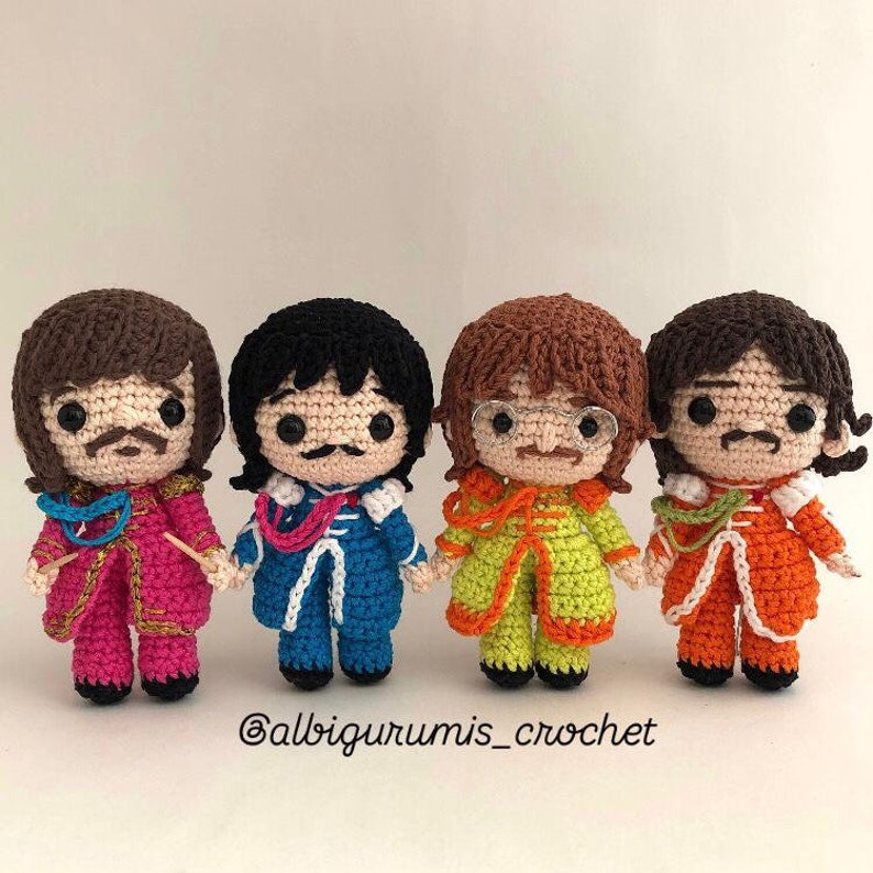 May include: Four crocheted dolls, each representing a member of the Beatles. The dolls are wearing colorful outfits and have detailed facial features. The dolls are standing in a row on a white surface. The caption at the bottom of the image reads "@albigurumis_crochet".