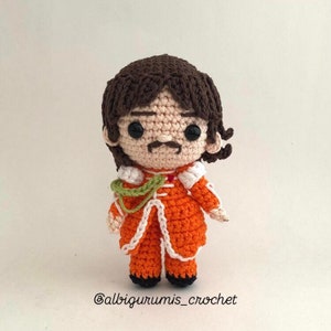 May include: Crocheted doll with brown hair, a mustache, and wearing an orange and white outfit. The doll is standing on a white background. The text '@albigurumis_crochet' is at the bottom of the image.