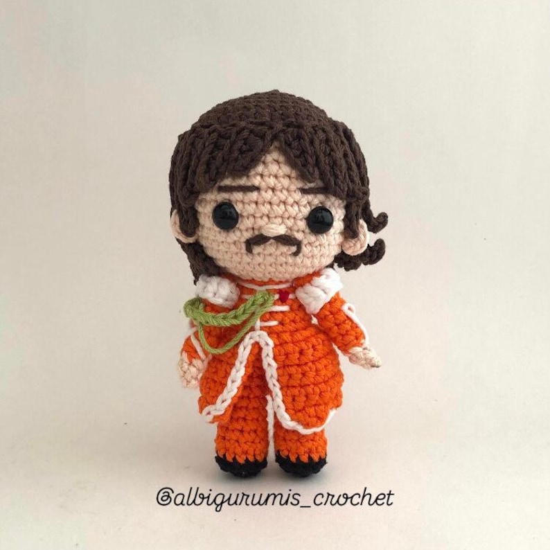 May include: Crocheted doll with brown hair, a mustache, and wearing an orange and white outfit. The doll is standing on a white background. The text '@albigurumis_crochet' is at the bottom of the image.