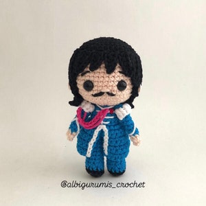 May include: A crocheted amigurumi doll with black hair, a blue suit, a pink scarf, and a mustache. The doll has large black eyes and a white face. The doll is standing on a white background. @albigurumis_crochet