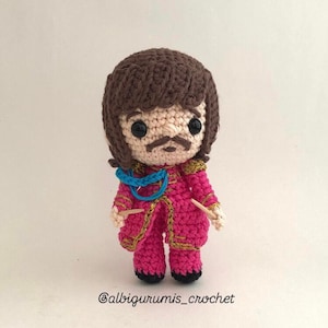 May include: Crocheted amigurumi doll with brown hair, a pink jacket with gold trim, and a blue scarf. The doll is holding two wooden sticks.