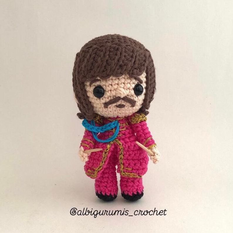 May include: Crocheted amigurumi doll with brown hair, a pink jacket with gold trim, and a blue scarf. The doll is holding two wooden sticks.