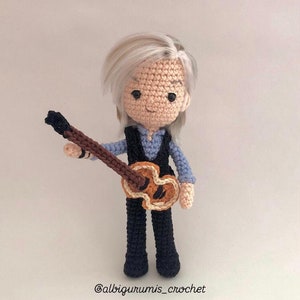 Op de afbeelding: Gehaakte amigurumi pop met wit haar, een blauw shirt, een zwart vest en een zwarte broek. De pop houdt een bruine en oranje gitaar vast.