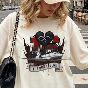 May include: Cream-coloured t-shirt with a graphic featuring a black heart, a jet, and the text "THE BIFF LEGEND". The shirt is paired with a beige baseball cap and a black purse with a chain strap. The image is taken outdoors.