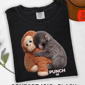 May include: Black Comfort Colors t-shirt featuring a graphic of a plush orangutan hugging a sleeping monkey. The word "PUNCH" is printed below the image. The text "COMFORT 1717 - BLACK" is printed at the bottom.