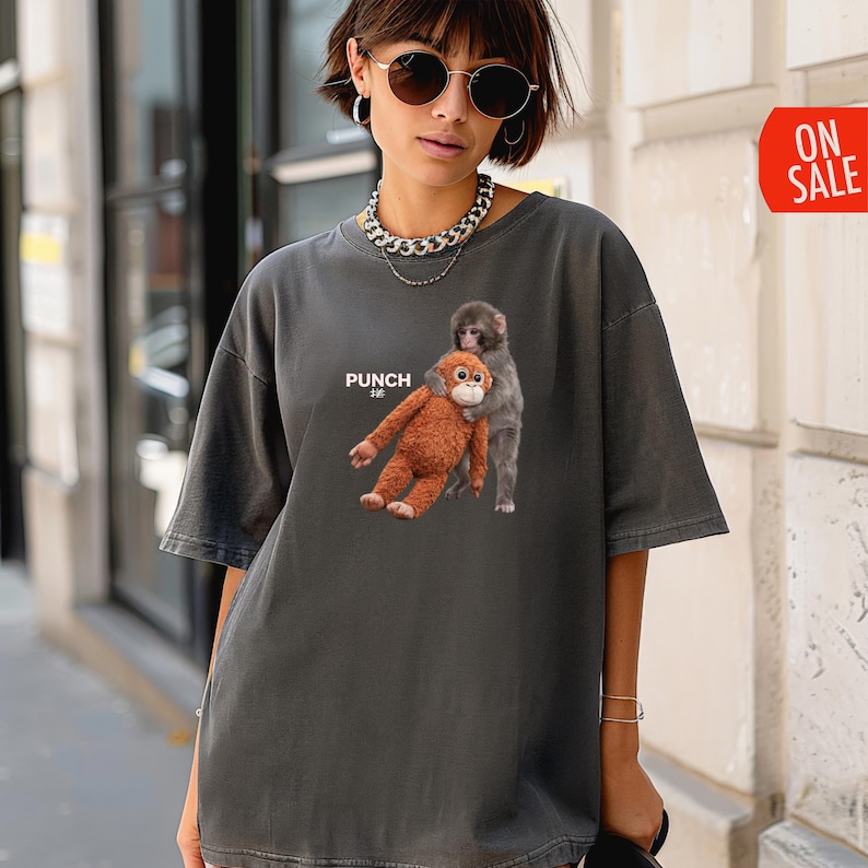 May include: Dark gray oversized t-shirt featuring a graphic of two monkeys. One is brown and the other is gray. The word "PUNCH" is printed on the shirt. The shirt has short sleeves and a relaxed fit.