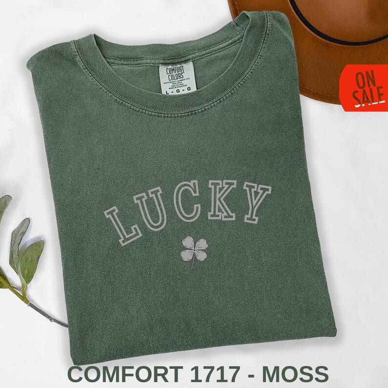 A flat lay of a folded Comfort Colors t-shirt in Moss Green. The design features the word 'LUCKY' in a retro, white collegiate-style arched font with a minimalist four-leaf clover icon underneath. The shirt is accessorized with a brown hat and a small green leaf branch on a white background, highlighting a vintage Saint Patrick's Day aesthetic