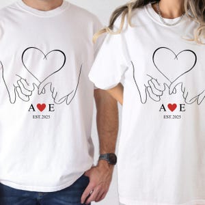 Op de afbeelding: Twee witte T-shirts met een zwarte lijntekening van twee handen die een hart vasthouden. De tekst "A ♥ E EST. 2025" is onder het hart gedrukt.