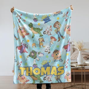 May include: A light blue blanket featuring various Toy Story characters, including Woody, Buzz Lightyear, and Mr. Potato Head. The name "Thomas" is displayed in large yellow letters. The blanket is held up by a person.