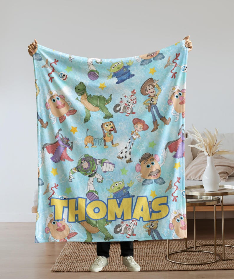 May include: A light blue blanket featuring various Toy Story characters, including Woody, Buzz Lightyear, and Mr. Potato Head. The name "Thomas" is displayed in large yellow letters. The blanket is held up by a person.