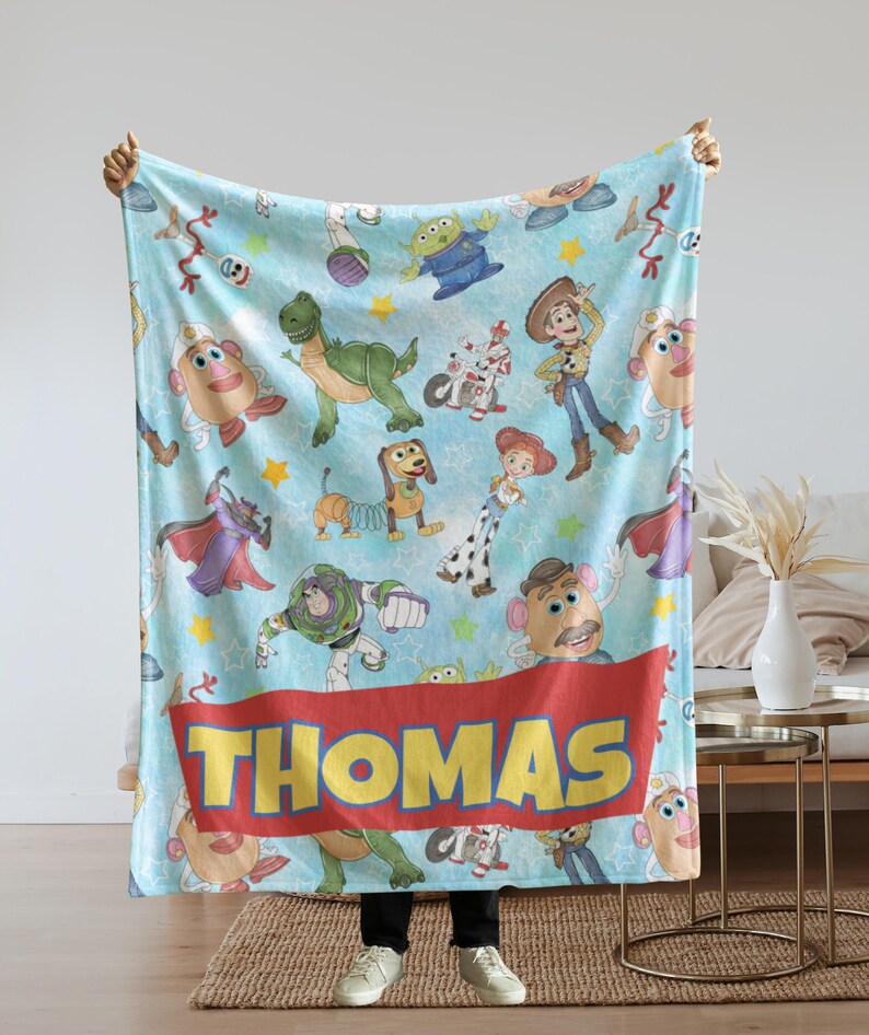 May include: A light blue blanket featuring various Toy Story characters, including Woody, Buzz Lightyear, and Mr. Potato Head. The blanket has a red banner with the name "THOMAS" in yellow letters. The blanket is held up by a person.