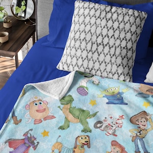 May include: A light blue blanket featuring various Toy Story characters, including Mr. Potato Head, Rex, Buzz Lightyear, and Woody. The blanket has a white sherpa backing and is placed on a blue bed with a patterned pillow.