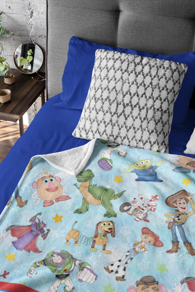 May include: A light blue blanket featuring various Toy Story characters, including Mr. Potato Head, Rex, Buzz Lightyear, and Woody. The blanket has a white sherpa backing and is placed on a blue bed with a patterned pillow.