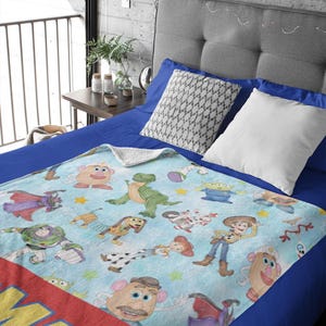 May include: A light blue blanket featuring various Toy Story characters, including Woody, Buzz Lightyear, and Mr. Potato Head. The blanket has a red border with the word "MAS" in yellow. The blanket is on a blue bed.
