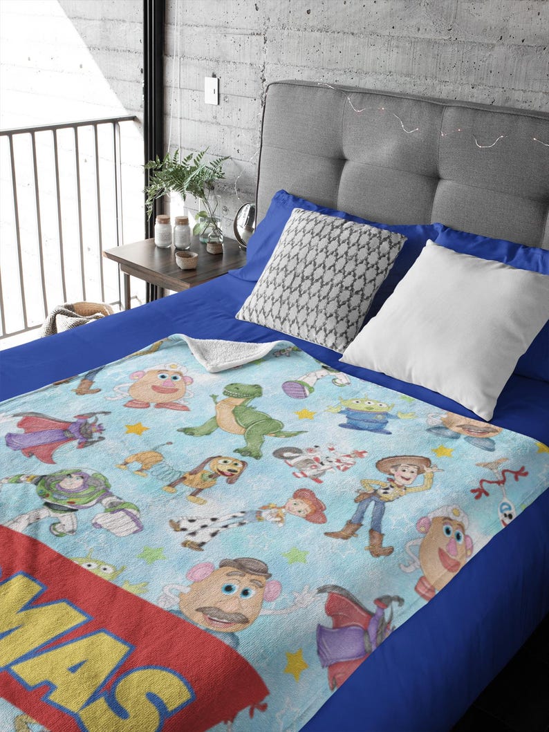 May include: A light blue blanket featuring various Toy Story characters, including Woody, Buzz Lightyear, and Mr. Potato Head. The blanket has a red border with the word "MAS" in yellow. The blanket is on a blue bed.