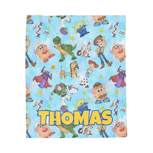 May include: A light blue blanket featuring a repeating pattern of Toy Story characters, including Woody, Buzz Lightyear, and Mr. Potato Head. The name "THOMAS" is displayed in large yellow letters across the bottom. The blanket is a soft, cozy material.