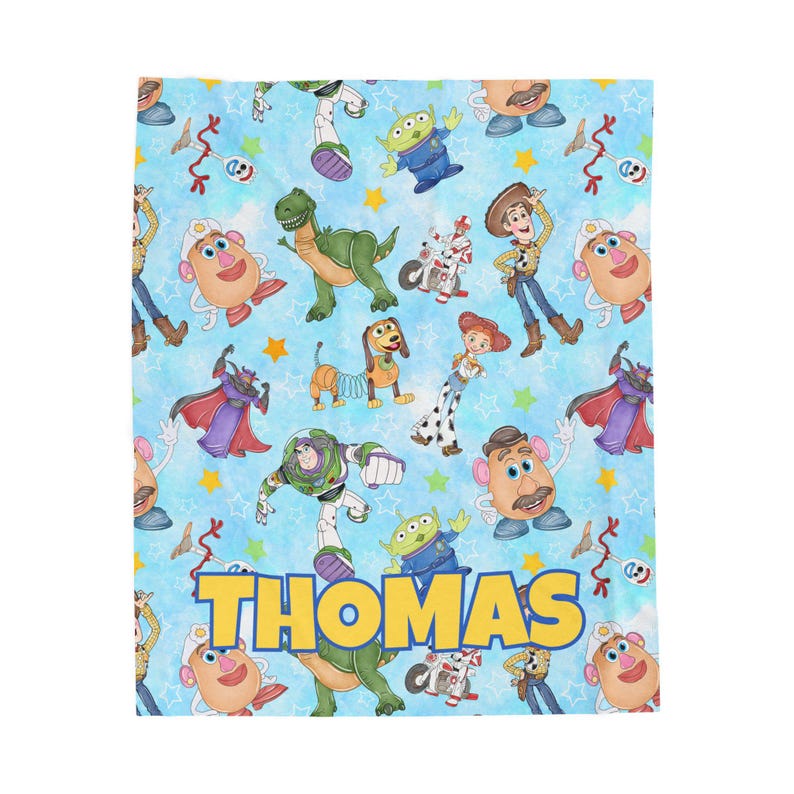 May include: A light blue blanket featuring a repeating pattern of Toy Story characters, including Woody, Buzz Lightyear, and Mr. Potato Head. The name "THOMAS" is displayed in large yellow letters across the bottom. The blanket is a soft, cozy material.