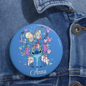May include: A round, blue button featuring the character Stitch from Lilo & Stitch. Stitch is depicted in a seated position in front of a castle. The button has a bow and Mickey Mouse-shaped balloons. The name "Anna" is written in cursive.