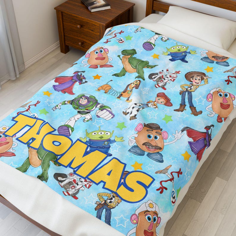 May include: A light blue blanket featuring various Toy Story characters, including Buzz Lightyear, Woody, and Mr. Potato Head. The name "THOMAS" is prominently displayed in large yellow letters. The blanket is decorated with stars and other playful elements.