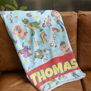 May include: A light blue blanket featuring various Toy Story characters, including Woody, Buzz Lightyear, and Rex. The blanket has a red band with the name "THOMAS" in yellow letters. The blanket is made of a soft material.