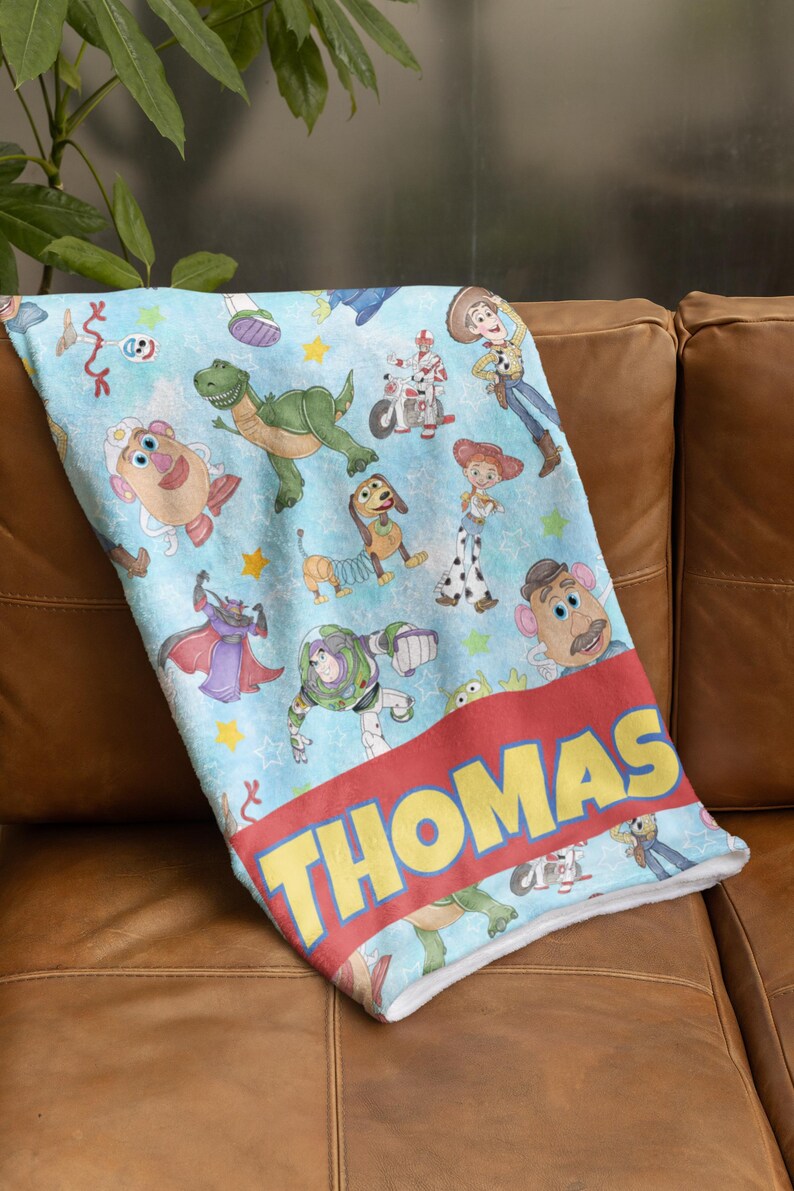 May include: A light blue blanket featuring various Toy Story characters, including Woody, Buzz Lightyear, and Rex. The blanket has a red band with the name "THOMAS" in yellow letters. The blanket is made of a soft material.