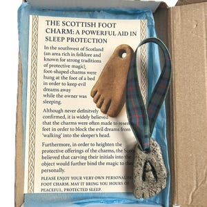 May include: A Scottish foot charm presented in a cardboard box. The charm, shaped like a foot, has the letter "A" carved into it and hangs from a green and blue tartan ribbon. The box includes text describing the charm's use.