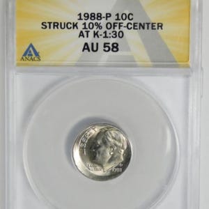 May include: A silver 1988-P 10C coin, graded AU 58, encased in a clear plastic holder. The coin is off-center and features the text "1988-P 10C STRUCK 10% OFF-CENTER AT K-1:30 AU 58" on a yellow and white label. The coin is a dime.