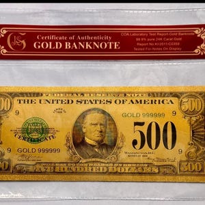 May include: A gold banknote, encased in a clear protective sleeve, with a red certificate of authenticity. The note features the text "THE UNITED STATES OF AMERICA" and the denomination "500". The certificate reads "GOLD BANKNOTE".