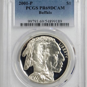May include: A silver 2001-P Buffalo commemorative dollar coin encapsulated in a clear plastic PCGS slab. The coin features a profile of a Native American with the word LIBERTY and the year 2001. The label reads 2001-P $1 PCGS PR69DCAM Buffalo 99793.69/54899189.