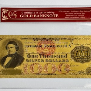 May include: A gold-plated $1,000 banknote with a portrait of a man in a suit and tie. The banknote is framed in a clear plastic sleeve with a red certificate of authenticity that reads "Certificate of Authenticity GOLD BANKNOTE" and "COA Laboratory Test Report-Gold Banknote 99.9% pure 24K Carat Gold Report No:K1/2011/C0369 Tested For-Notes On Display".