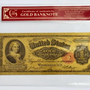 May include: A gold-colored $1 United States banknote with a portrait of a man. The banknote is framed in a clear plastic sleeve with a red certificate of authenticity that reads "Gold Banknote".