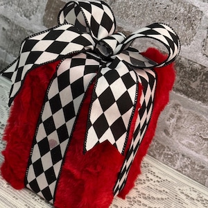 May include: A square-shaped decorative gift box covered in bright red faux fur. The box is wrapped with a wide ribbon featuring a black and white diamond harlequin pattern, tied into a large, multi-looped bow on the top center of the box.