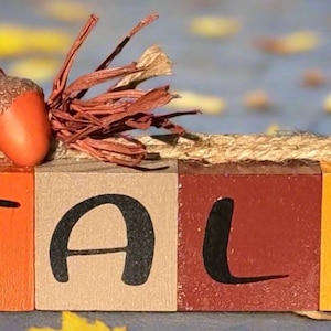 May include: A wooden block sign spelling "FALL" in black letters. The blocks are painted in orange, tan, red, and yellow. An acorn and decorative twigs sit atop the sign, which is wrapped with twine. The background is blurred with fall leaves.