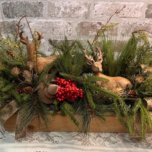 May include: A rectangular wooden box centerpiece featuring two brown deer figurines nestled among artificial pine branches, green foliage, small pinecones, and clusters of bright red berries, accented with a patterned tan fabric ribbon.