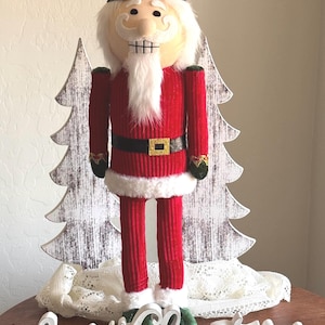 May include: A nutcracker figure wearing a dark brown hat with a gold emblem, a red jacket with a black belt, and white faux fur trim. Behind the figure are two white wooden tree cutouts, and in front is a white sign that reads Merry Christmas.