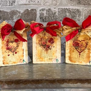 May include: Three rectangular wooden plaques featuring vintage-style floral heart illustrations, aged paper textures, and handwritten script. Each plaque is topped with a large red satin bow and a beige lace ribbon, with letters A, B, and C marked.