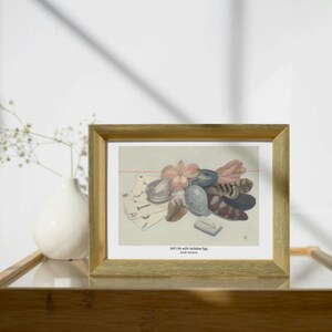 Puede incluir: Una impresión enmarcada titulada "Still Life with Jackdaw Egg" de Sarah Homan. La obra presenta un arreglo de naturaleza muerta de objetos, incluyendo un huevo, naipes y flores. El marco es dorado.