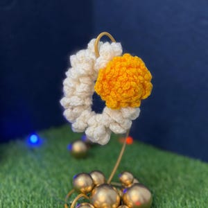 Può includere: Un elastico per capelli in crochet bianco e giallo su un supporto in metallo dorato. L'elastico è fatto di filato morbido e ha un design a fiore.