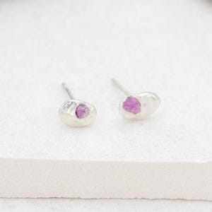 May include: Two silver stud earrings, each featuring a small, pink gemstone. The earrings are presented on a white surface, highlighting their minimalist design. The gemstones have a raw, natural look, adding to their charm.